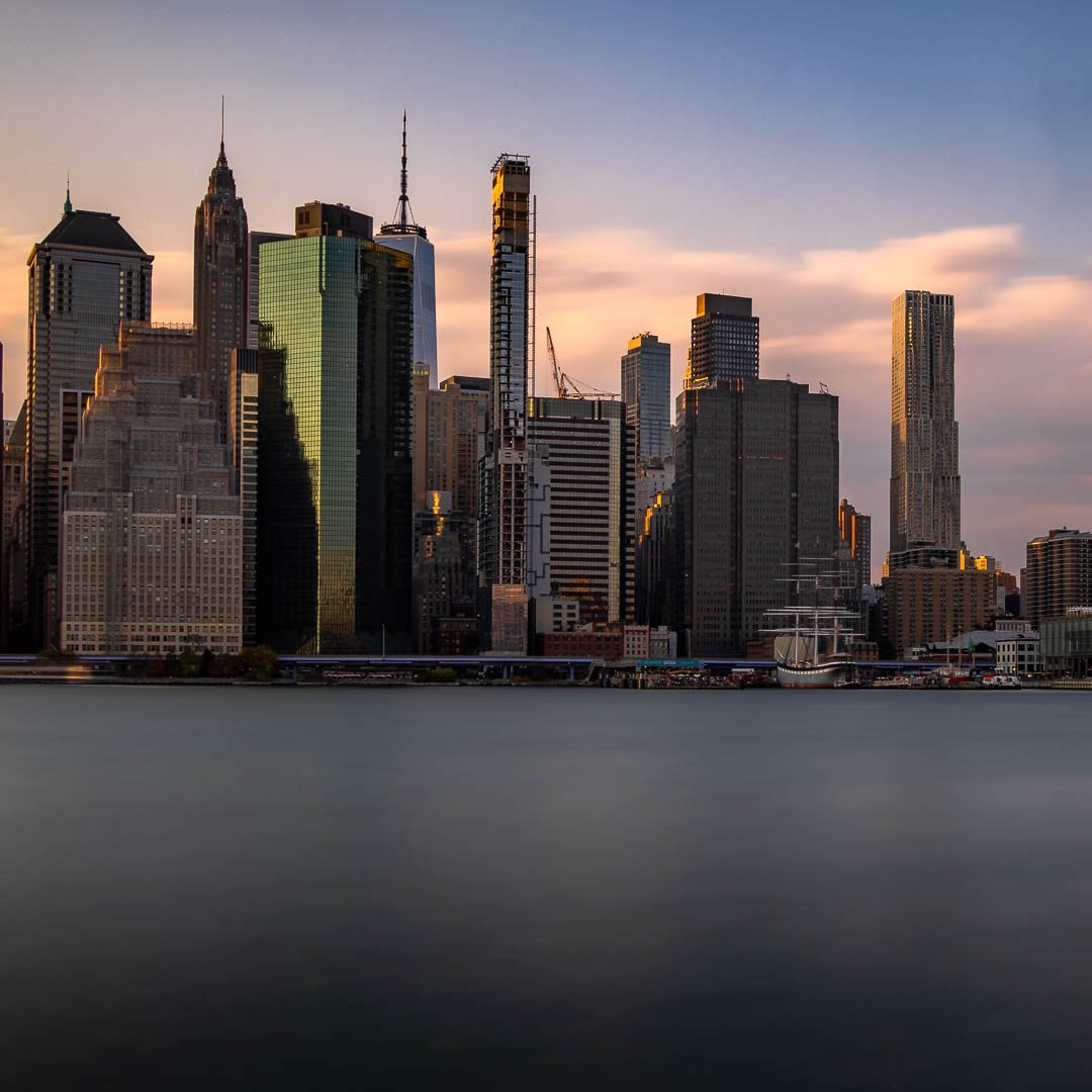 Skyline of Manhattan at sunset, tall buildings and calm waters in the foreground.