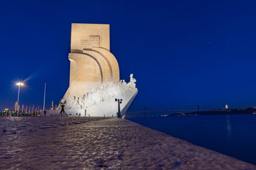 Monument with nave-like sculpture by the water, illuminated at night.