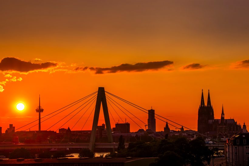 Sonnenuntergang über einer Stadt mit einer markanten Brücke und Türmen im Hintergrund.