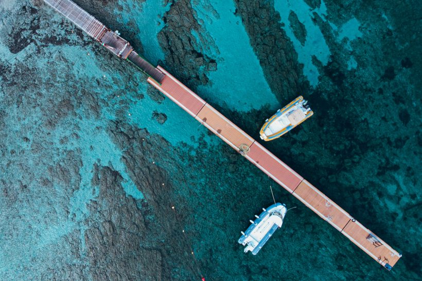 Two boats are located in shallow, turquoise waters next to a long, narrow footbridge.