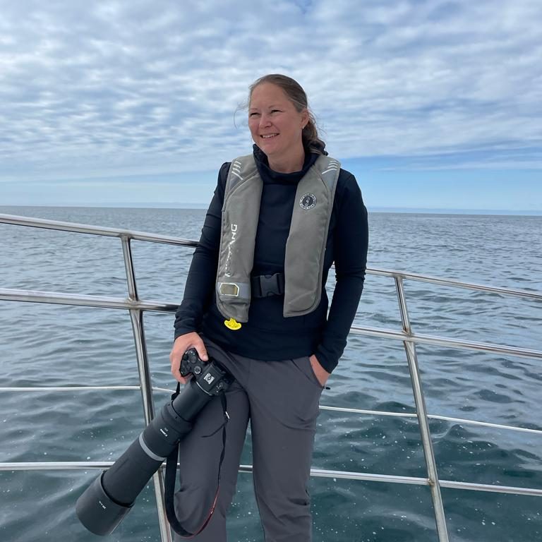 Woman with camera is standing on a boat, background with water and cloudy sky.