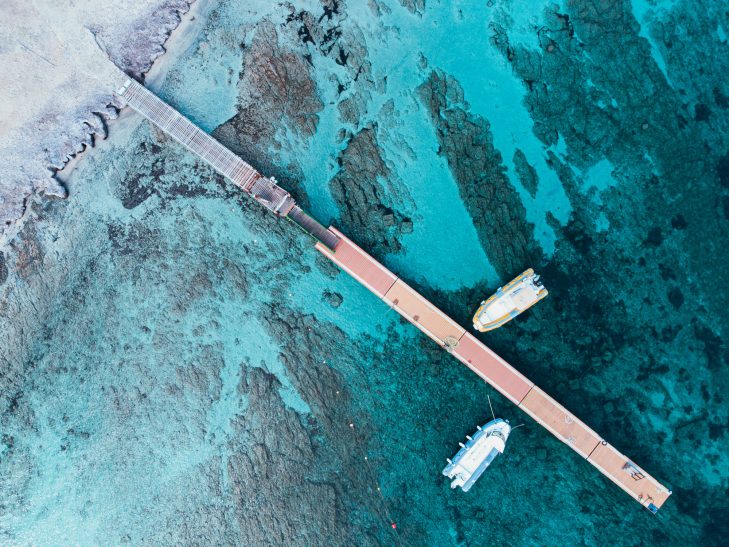 Eine Holzpier ragt ins klare, türkisfarbene Wasser, daneben liegen zwei Boote.