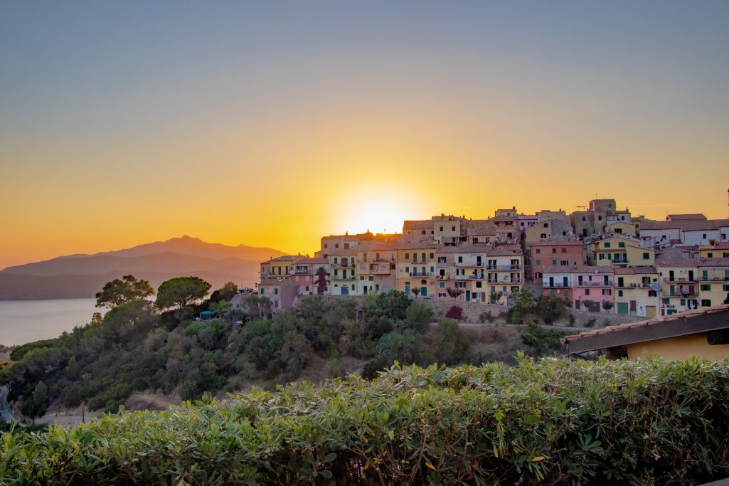 Sonnenuntergang über bunten Häusern an der Küste, mit Hügeln im Hintergrund.