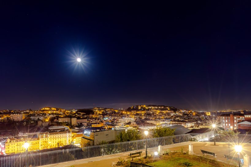 Nachtansicht einer Stadt mit beleuchteten Gebäuden und einem Vollmond am Himmel.