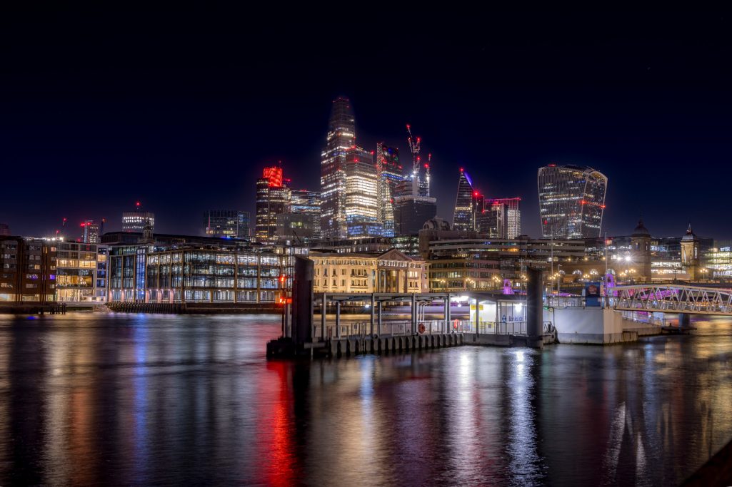 The skyline of London at night, illuminated with colorful lights and reflected in the water.