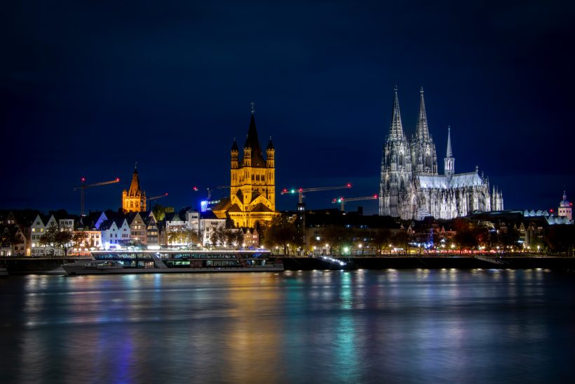 Cologne skyline at night with illuminated buildings and reflective water.