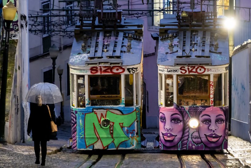 Two colorful trams with graffiti, one person with an umbrella walks past.