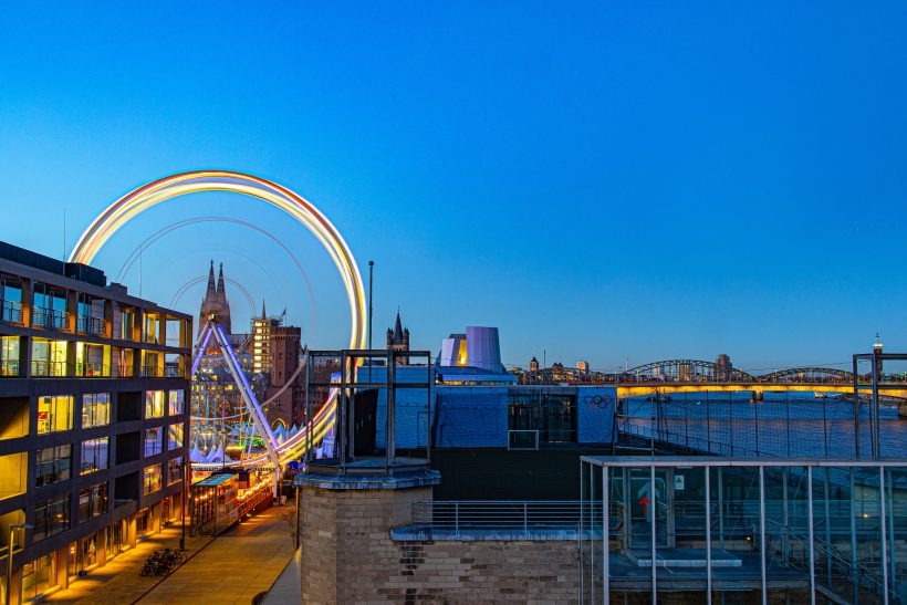 Riesenrad mit bunten Lichtern vor einem klaren blauen Himmel und Stadtansicht.