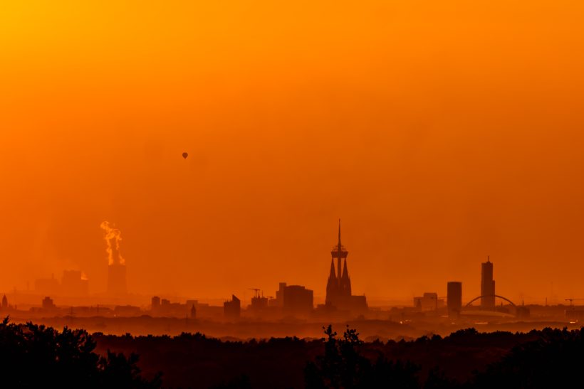 Silhouette einer Stadt bei Sonnenuntergang mit orangefarbenem Himmel und rauchenden Schornsteinen.