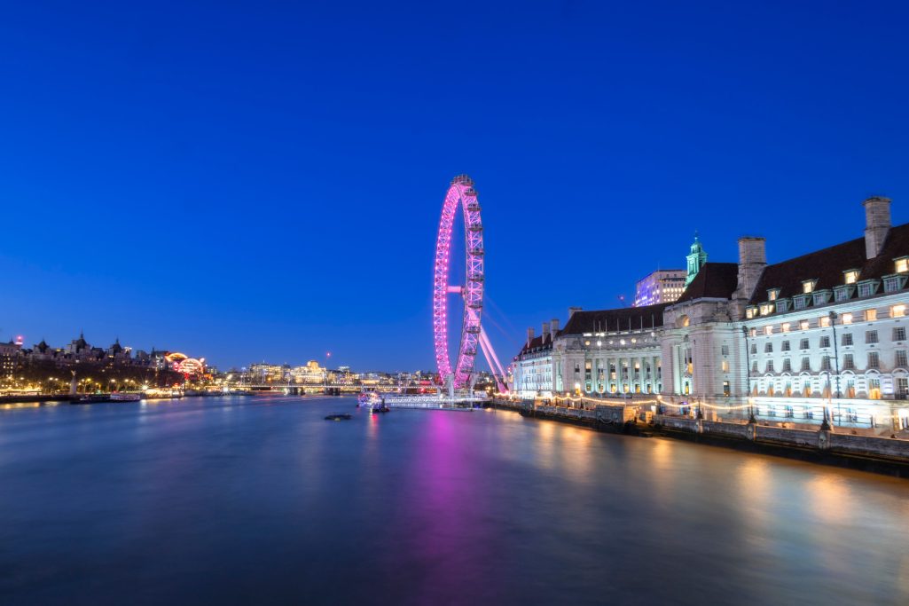 The London Eye shines in pink light in the evening over the Thames.