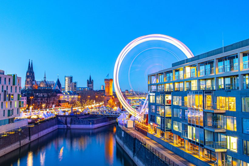 Nachtaufnahme der Stadt mit einem großen Riesenrad und beleuchteten Gebäuden entlang des Wassers.