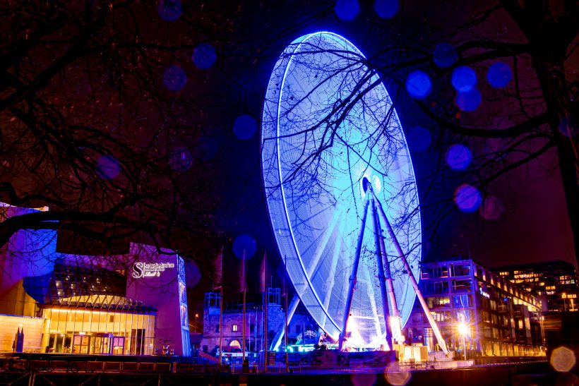 Riesenrad bei Nacht, leuchtend in blauen Lichtern, umgeben von Bäumen.