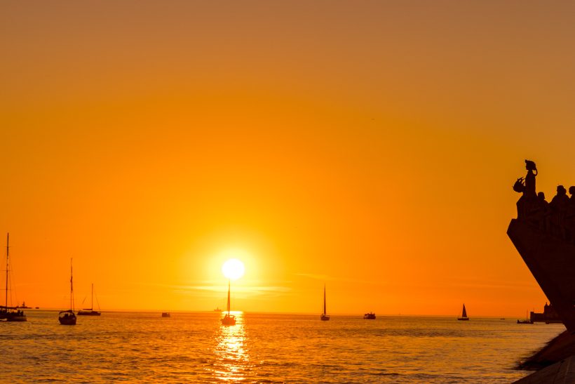 Sunset over the sea with sailboats and sharp silhouettes in the foreground.