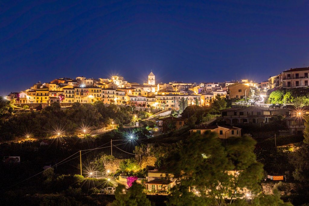 Night view of an illuminated city on a hill with surrounding landscape.