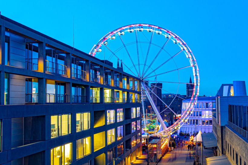 Ferris wheel with colorful lights in front of modern buildings at dusk.