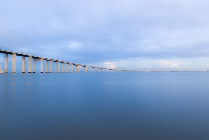 Eine lange Brücke mit Pfeilern über ruhiges Wasser unter einem bewölkten Himmel.