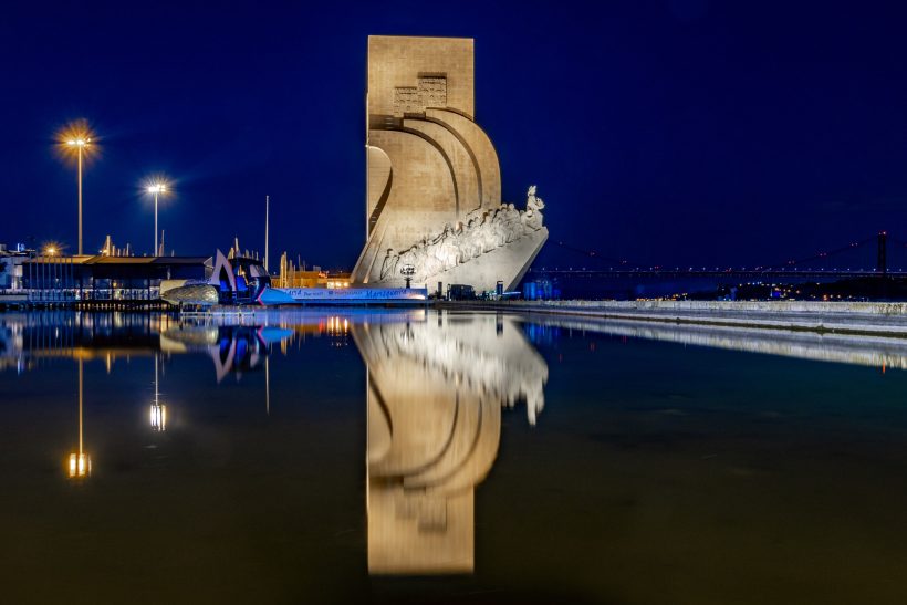 Monument mit gewundenen Formen, nachts beleuchtet, reflektiert im Wasser.