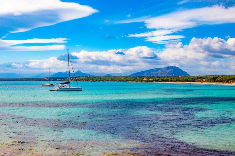 Blick auf turquoise Wasser mit Segelbooten und Bergen im Hintergrund unter blauem Himmel.