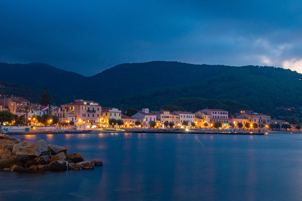 Dusk above a coastal village with illuminated houses and mountains in the background.