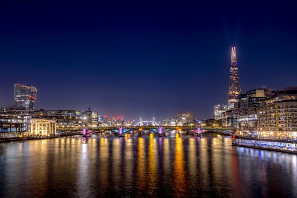 City view of London at night with illuminated buildings and a bridge over the river.