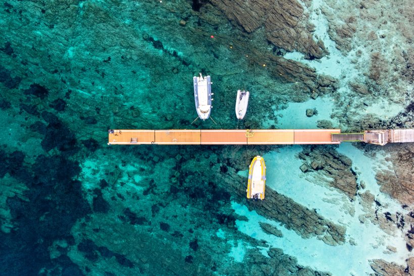 Wooden bridge over clear water with three boats and rocks in the background.