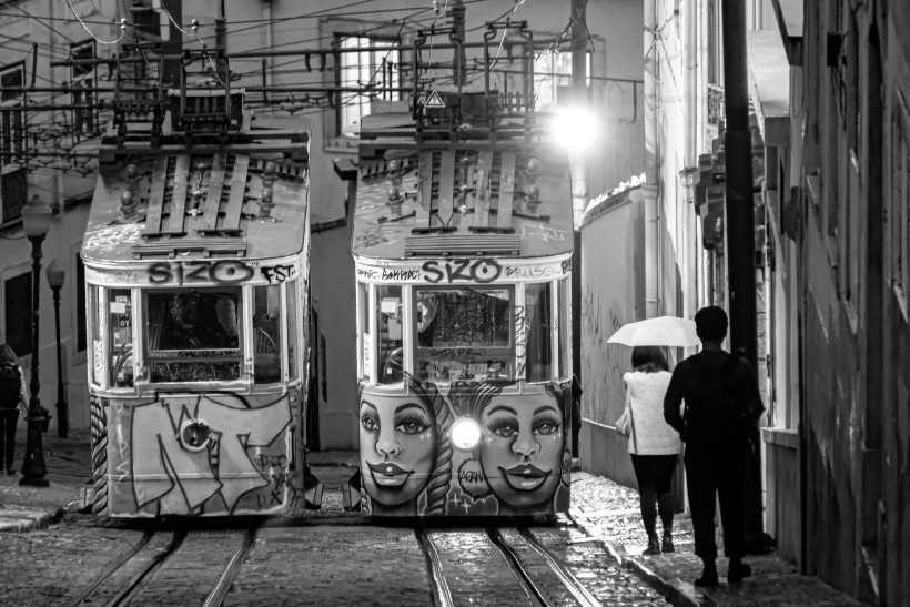 Zwei bemalte Straßenbahnen in einer engen Gasse bei Nacht, mit Personen und Regenschirm.