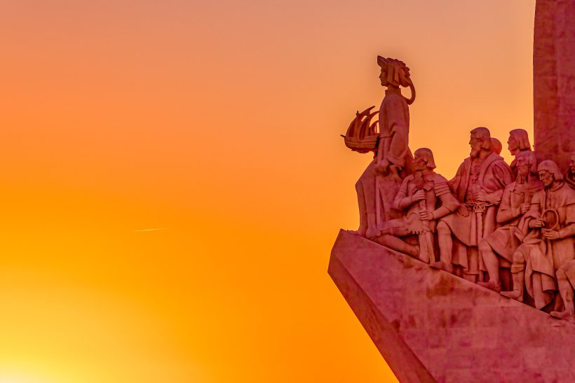 Statues of historical figures in front of an orange sunset.