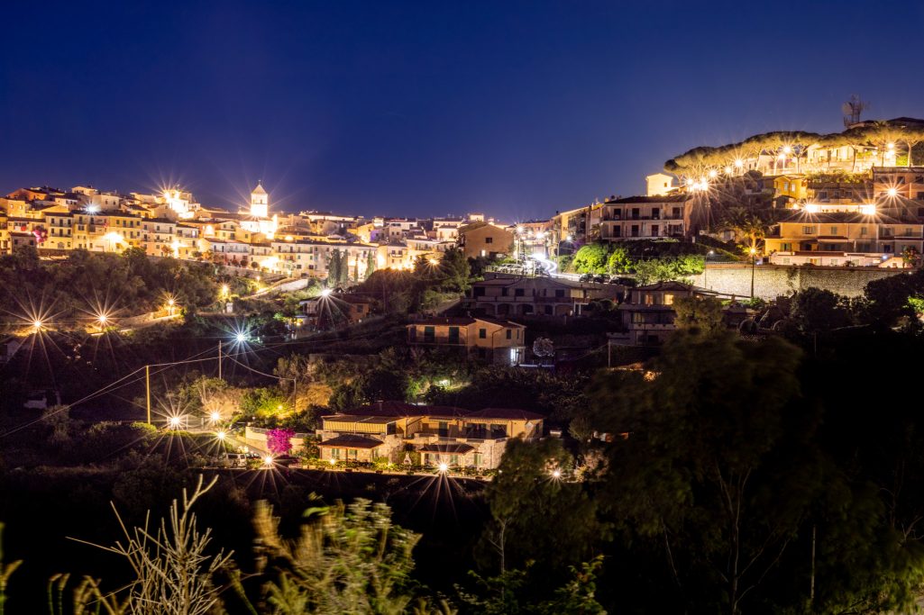 Nocturnal cityscape with illuminated buildings and rolling hills.