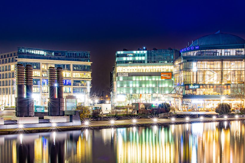 Modernes Gebäude entlang eines beleuchteten Ufers bei Nacht, mit Spiegelungen im Wasser.