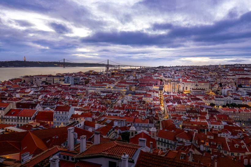 Blick auf die rote Dächersilhouette von Lissabon mit Himmel und Meer im Hintergrund.