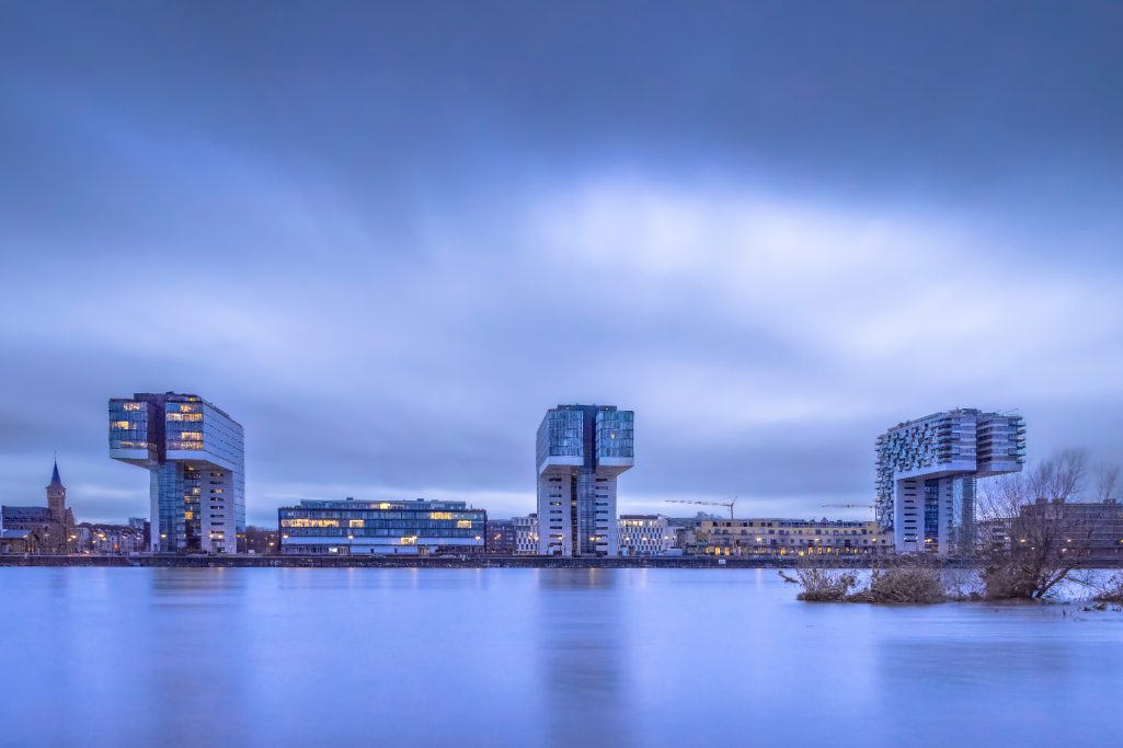 Three modern buildings on the water, surrounded by cloudy sky in cool blue.