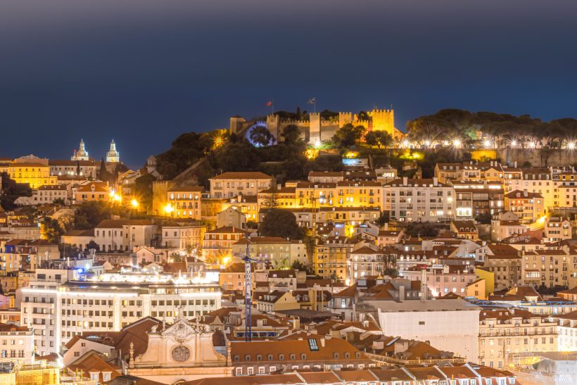 City view at night with illuminated buildings and a castle on a hill.