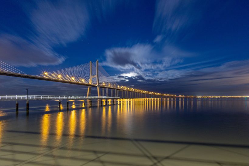 Illuminated bridge over calm water at dusk and dramatic sky.