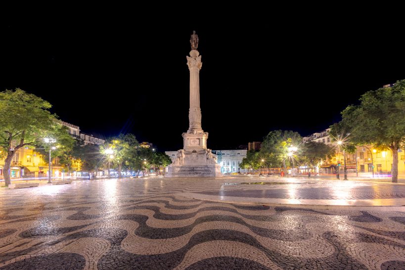 Square with a high monument and illuminated trees at night.