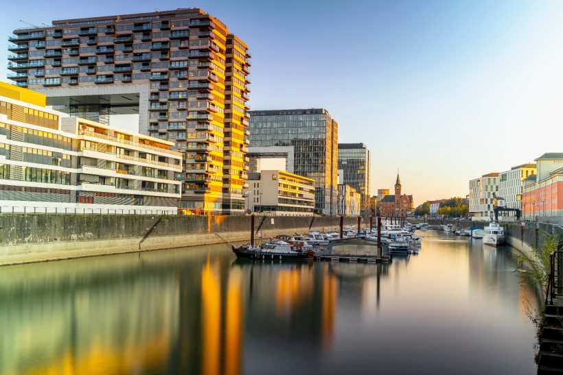 Modern buildings on the water with reflective surfaces and boats in the harbor.