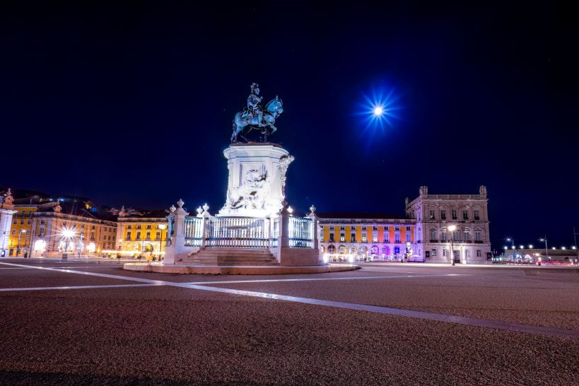 Eine beleuchtete Statue auf einem Platz bei Nacht, mit Mond und farbigem Hintergrund.