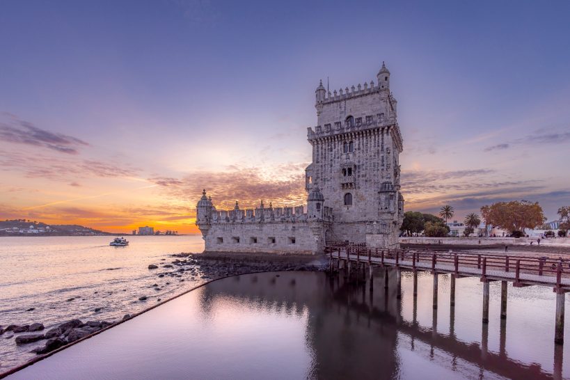 Torre de Belém on the water at sunset, with reflective colors in the foreground.