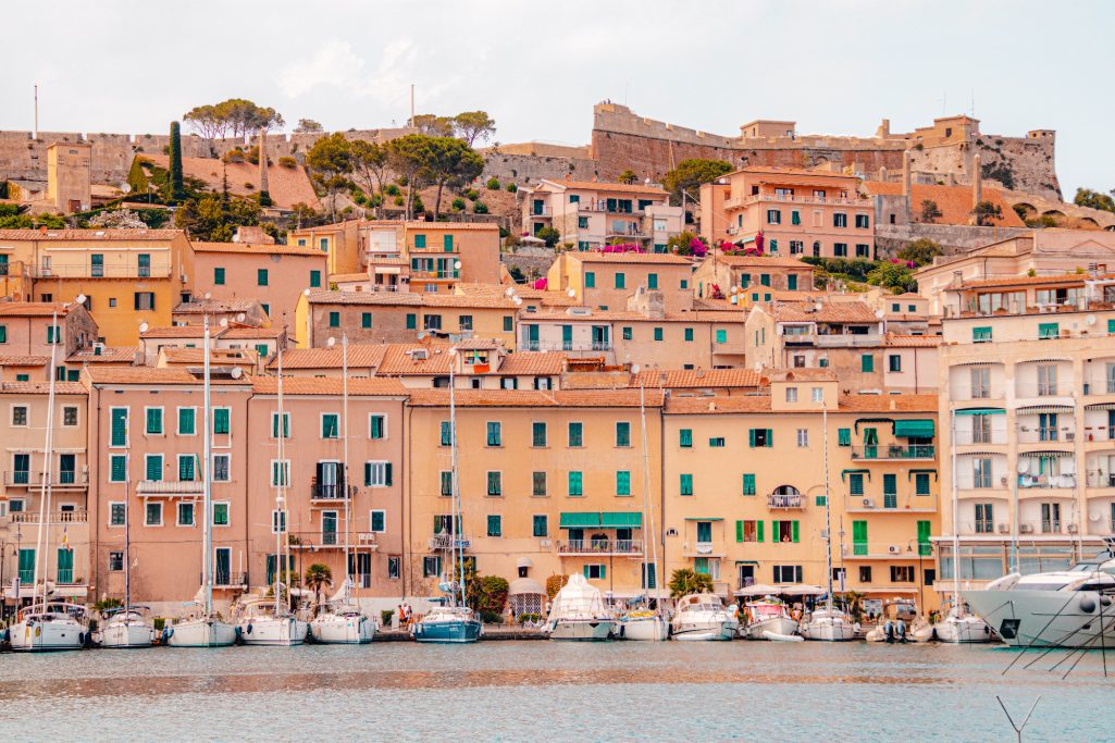 Colorful houses on the shore with sailboats and a historic fortress in the background.