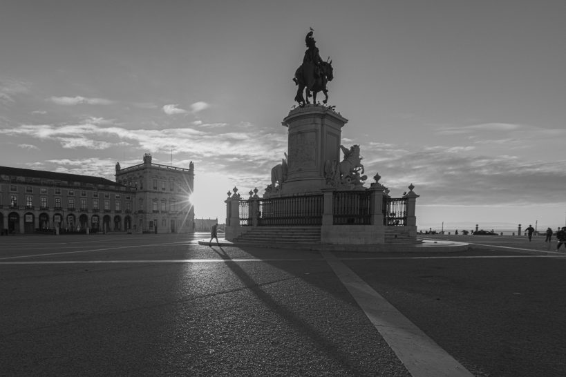 Sonnenaufgang hinter einer Statue auf einem Sockel, umgeben von Gebäuden.